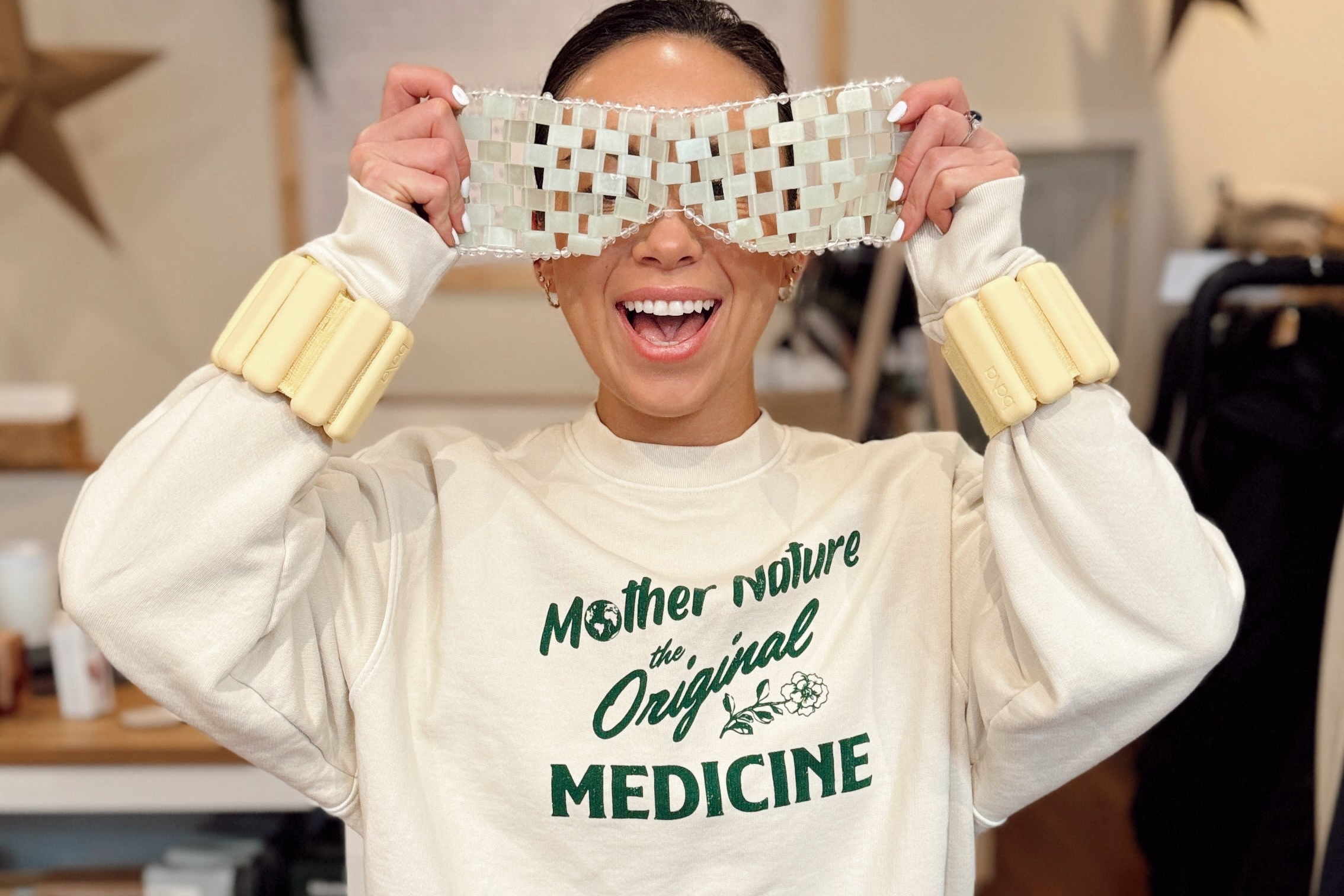 A smiling woman holding up a face mask with Bala Bangles around her wrists, wearing a sweatshirt that says "Mother Nature the Original Medicine"