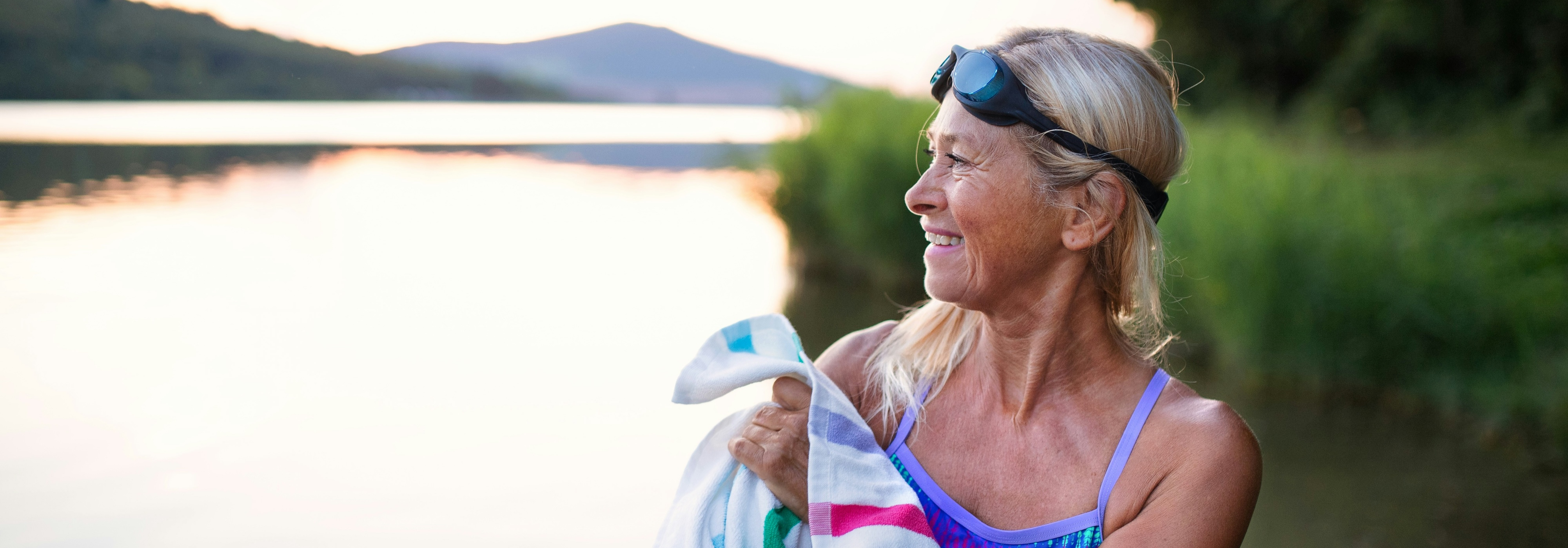 A woman in a blue swimsuit stands by a lake at sunset, holding a white towel with colorful stripes. She has black swim goggles on her head and is looking to the side.