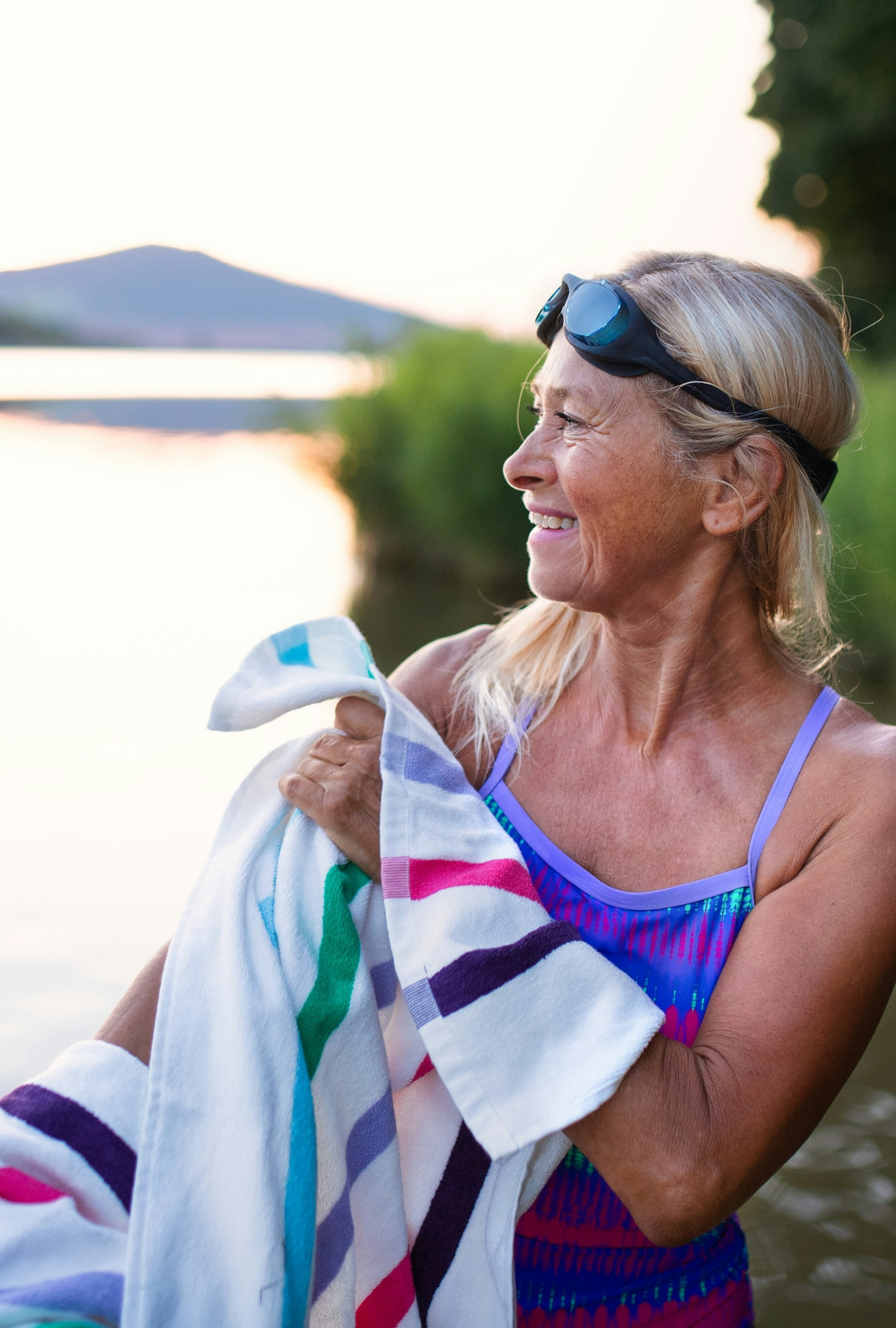 A smiling blonde woman outdoors by the water at sunset, wearing a blue swimsuit and holding a white towel with colorful stripes.