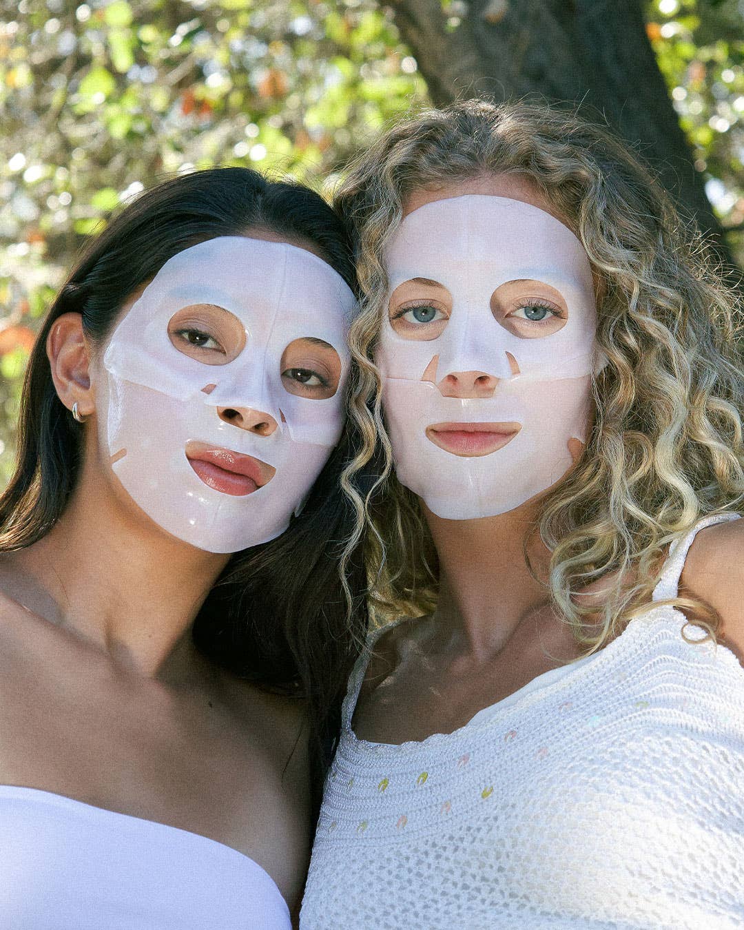 Two women wearing white face masks outdoors with greenery in the background