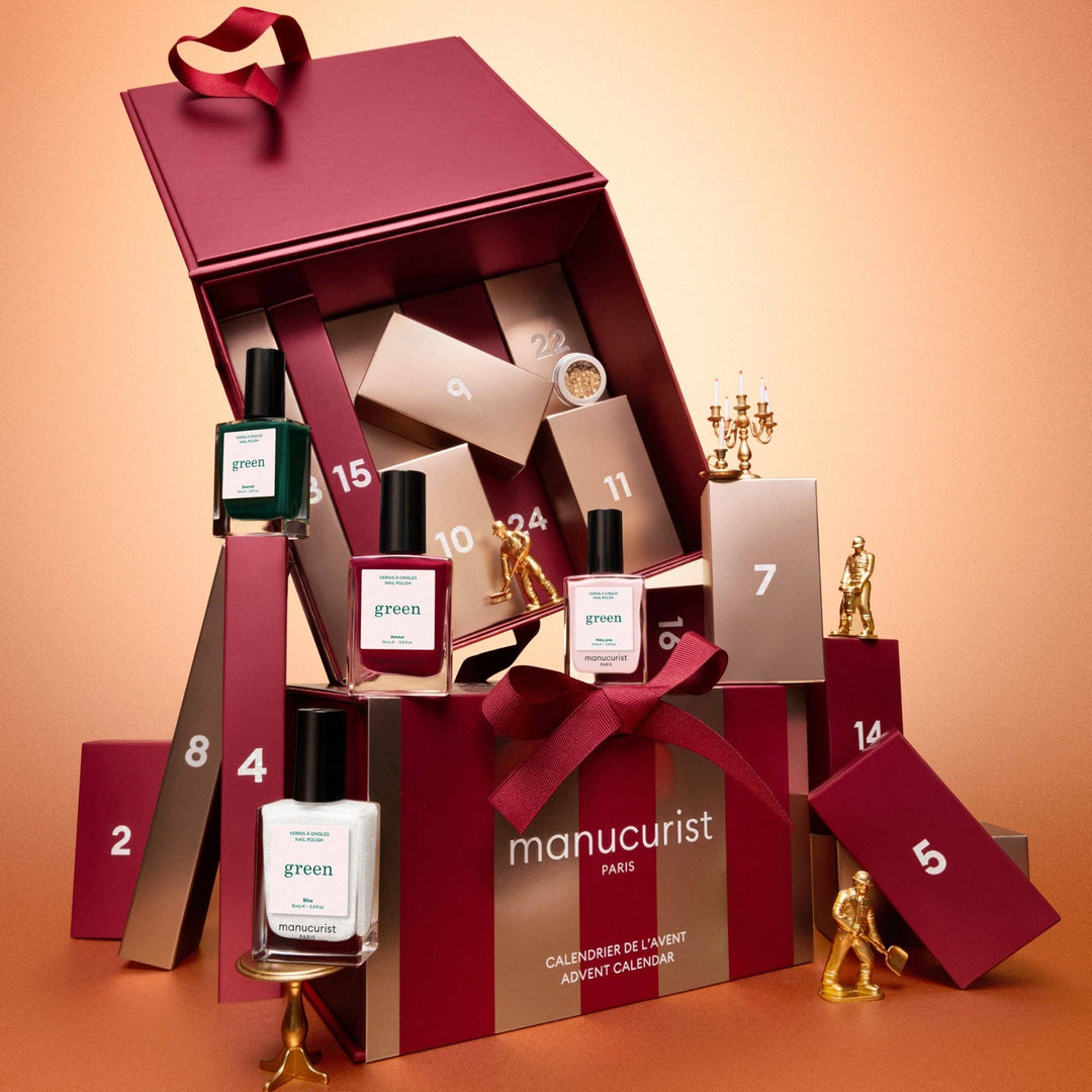 Manucurist Paris advent calendar gift set displayed open on a warm peach background, with numbered boxes, nail polish bottles, gold candlesticks, and a large burgundy gift box tied with a red ribbon.