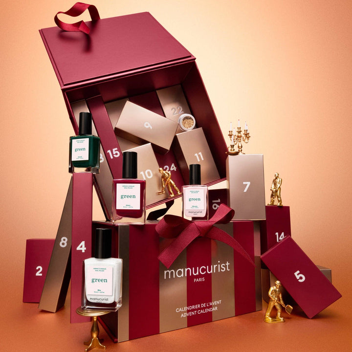 Manucurist Paris advent calendar gift set displayed open on a warm peach background, with numbered boxes, nail polish bottles, gold candlesticks, and a large burgundy gift box tied with a red ribbon.