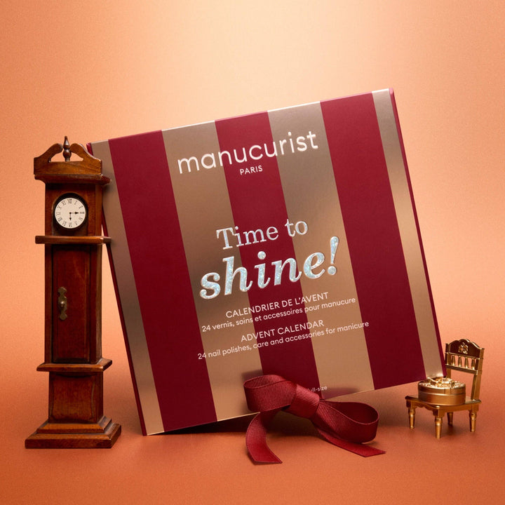 A manucurist Paris advent calendar package leaning against a warm peach background, styled with a miniature wooden grandfather clock on the left and a small gold vanity chair with a mirror on the right. A dark red ribbon is tied around the package at the bottom.