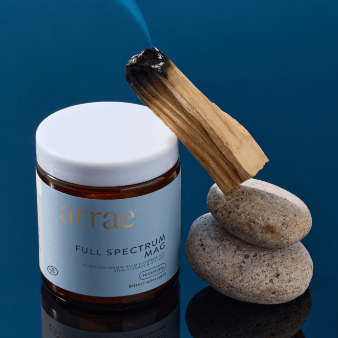 A jar of Arrae Full Spectrum Magnesium, shown next to a burning palo santo stick on stacked stones against a dark blue background.