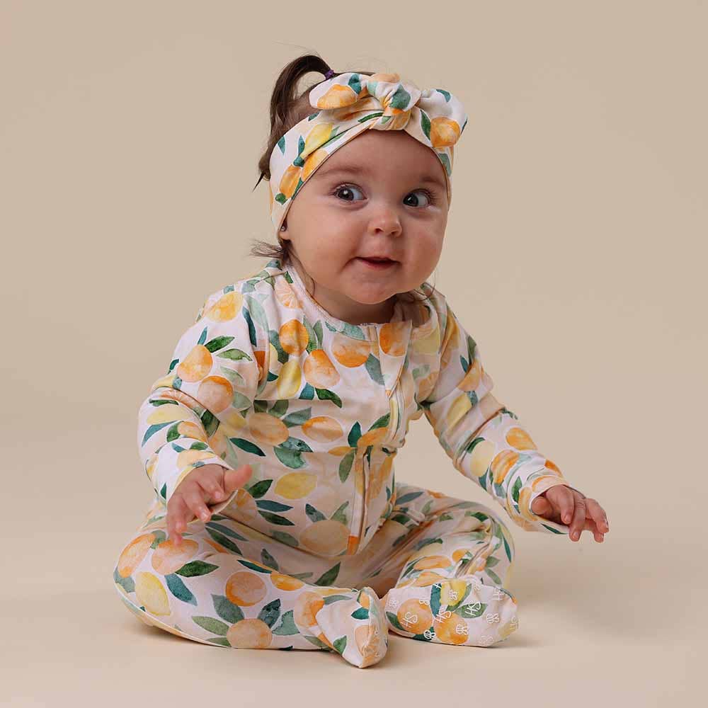 Baby wearing a Sunny Citrus Organic Snuggle Sleepsuit Zip Footie and headband on a beige background