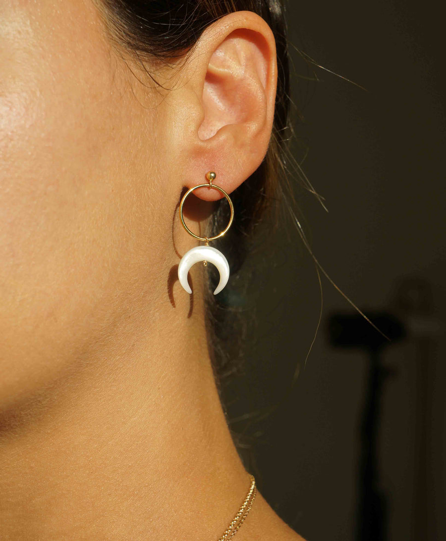 Close-up of an ear wearing a gold hoop earring with a white crescent moon charm against a dark background.