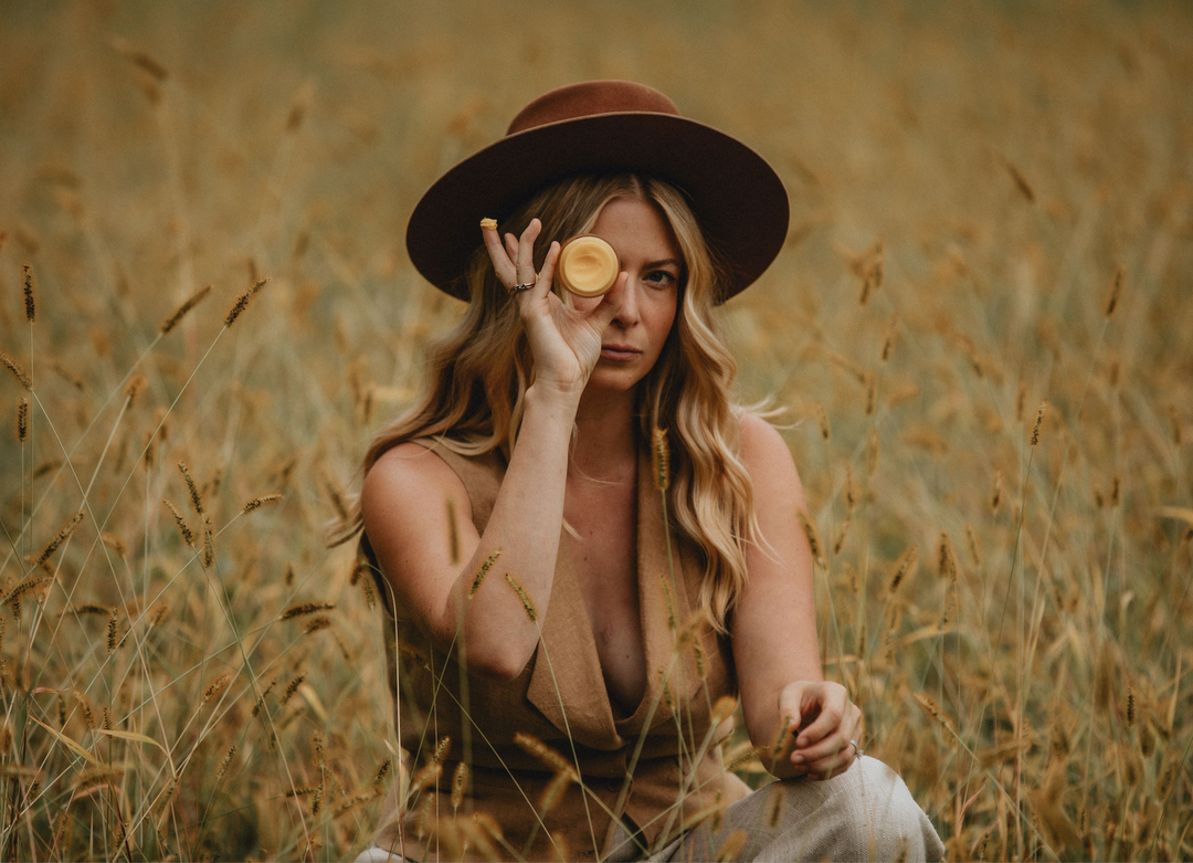 A woman in a field of tall grass wearing a hat and holding a jar of tallow.