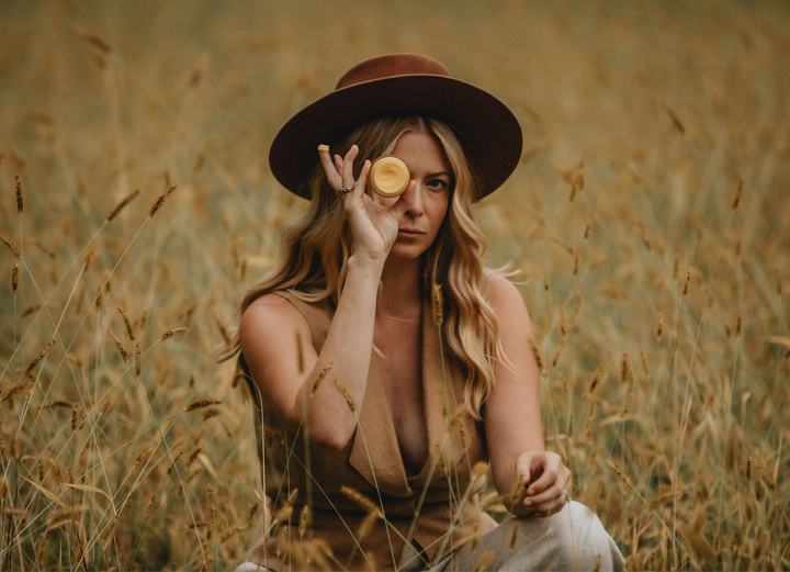 A woman in a field of tall grass wearing a hat and holding a jar of tallow.