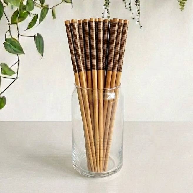 A set of bamboo chopsticks with dark brown striped handles, arranged in a clear glass jar on a light surface against a white wall, with green trailing leaves in the upper left corner.