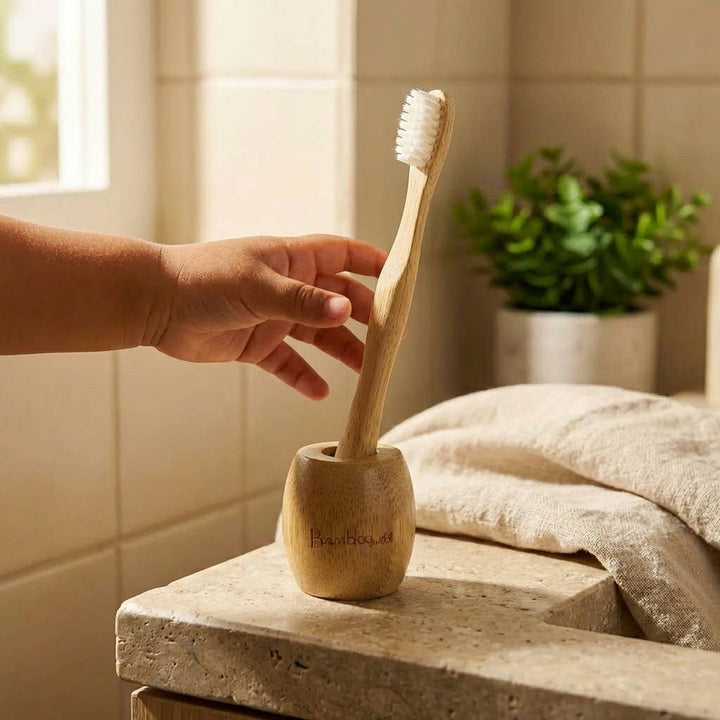 Hand holding a bamboo toothbrush over a wooden holder on a bathroom counter.