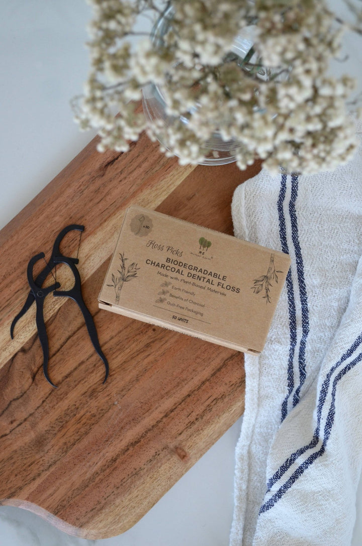Biodegradable charcoal dental floss packaging on a wooden board with a towel and flowers in the background.