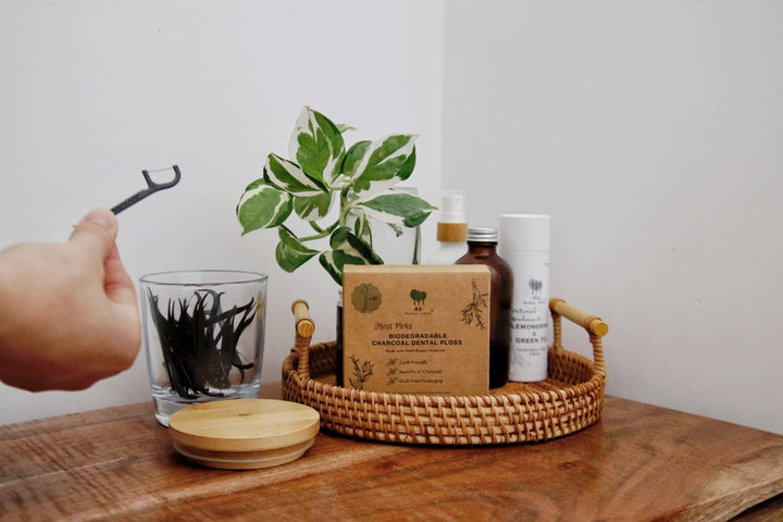 Hand holding a dental floss pick with a plant and various items on a wooden surface
