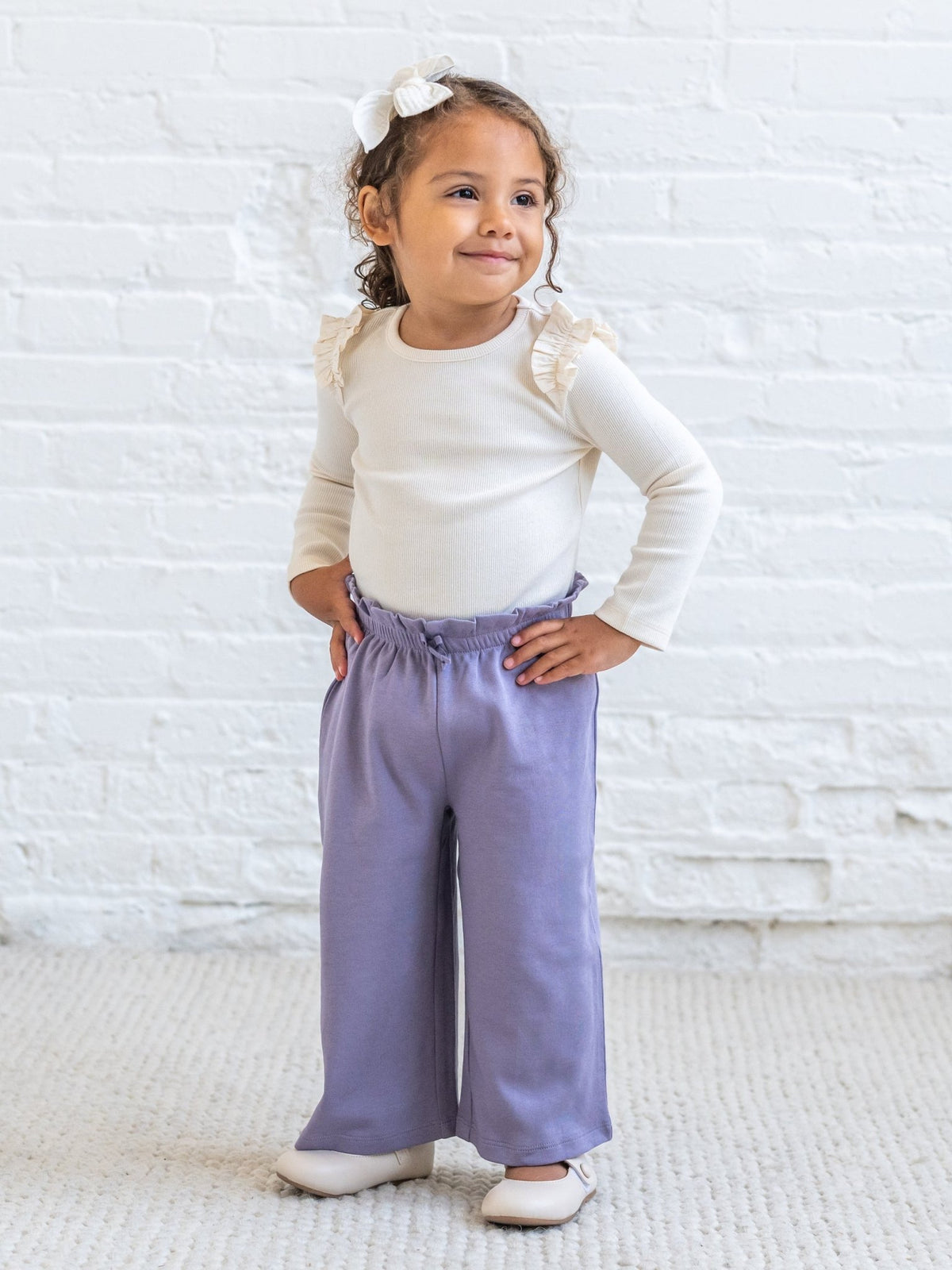 A young girl with a white bow in her hair stands with hands on hips, wearing a cream ribbed long-sleeve top with ruffle details on the shoulders and lavender wide-leg pants, set against a white brick wall background.