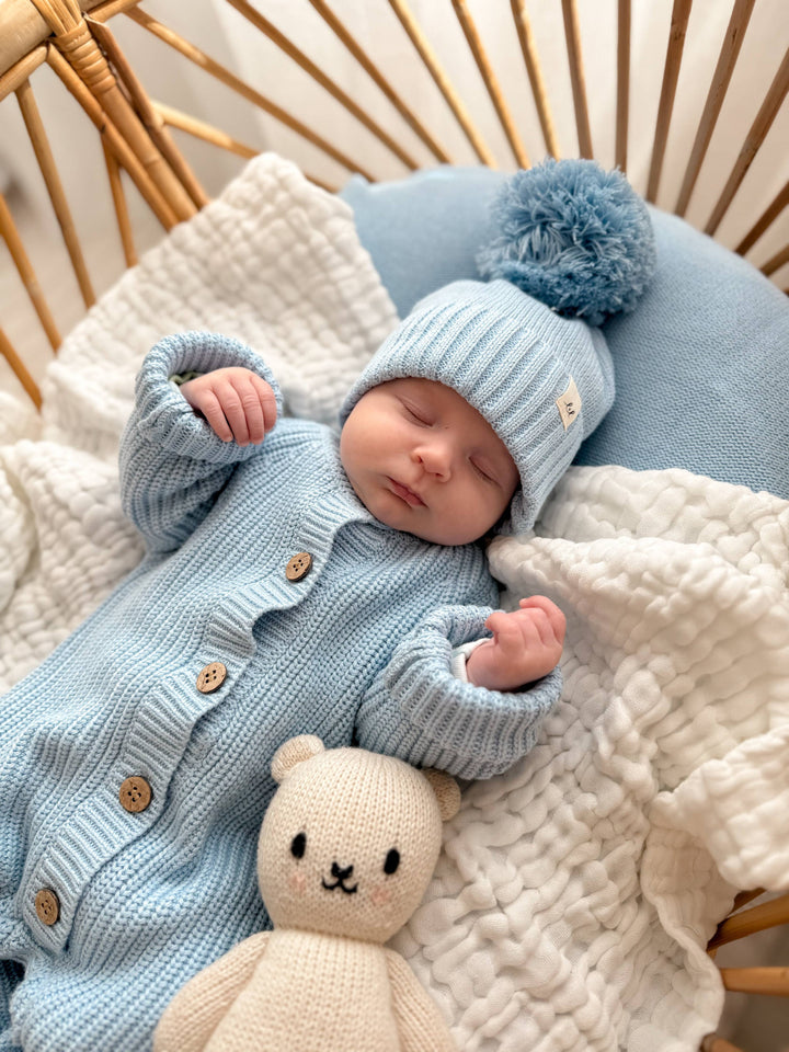 Newborn baby in a blue outfit and hat with a plush toy, lying on a textured blanket.