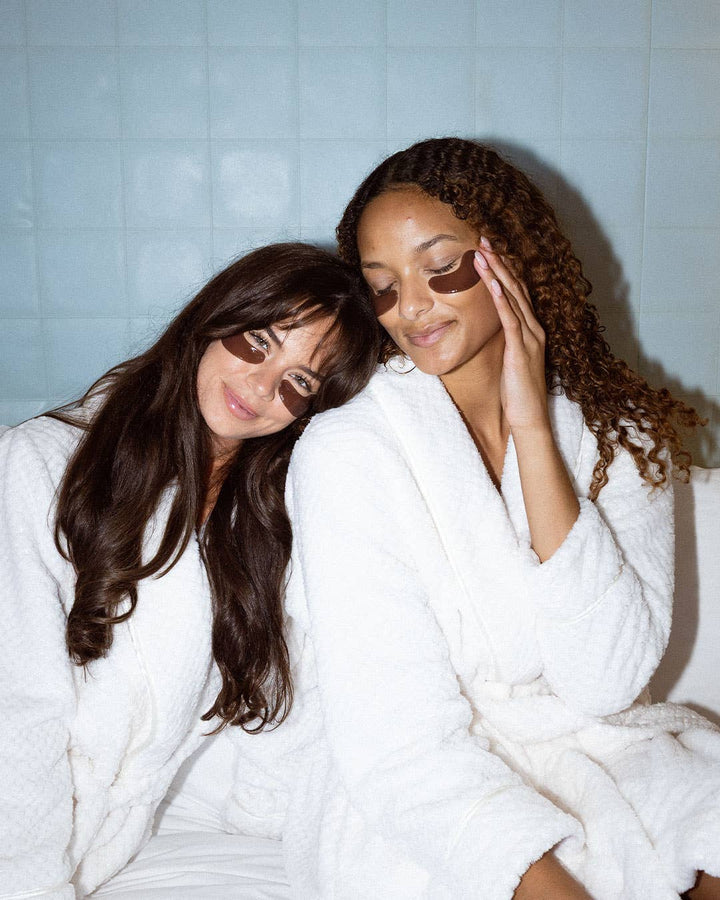 Two women in white robes with eye masks on a tiled wall background