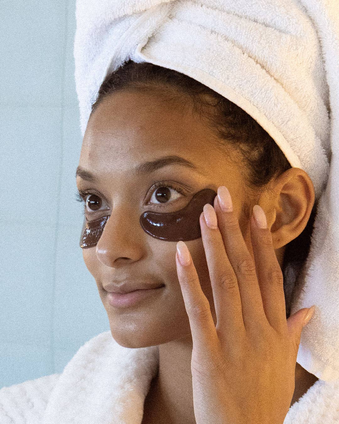 Woman with brown eye patches on her face, wearing a white towel, against a light blue background.