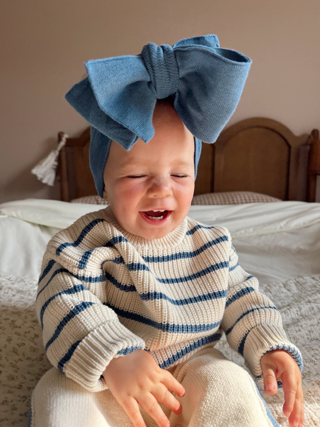 Baby wearing a blue striped sweater and large blue bow headband sitting on a bed.