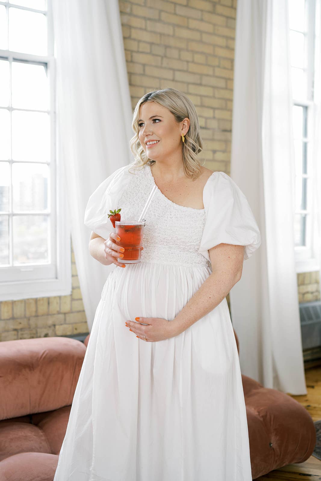 Pregnant woman in a white dress holding a tea in a room with large windows and a brick wall.