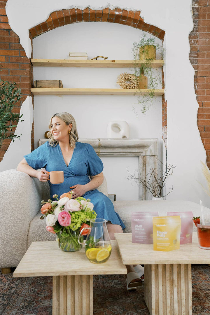 Woman in a blue dress sitting on a couch with a tea cup, flowers, and a pitcher of tea.