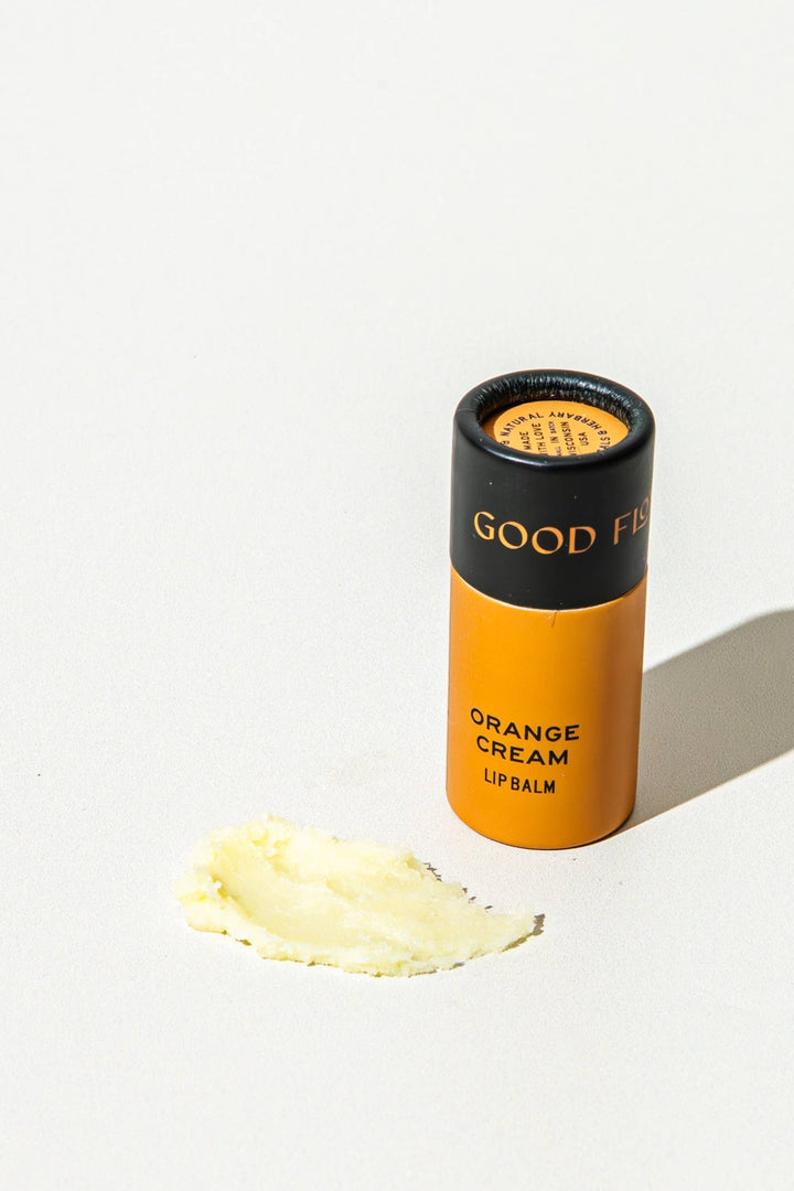 An orange and black tube of GOOD FLOWER FARM Orange Cream Lip balm stands on a white surface next to a smear of the pale yellow cream product.