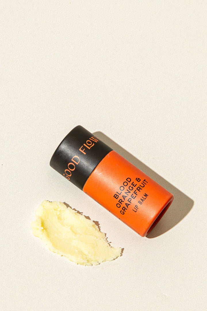A tube of Blood Orange &amp; Grapefruit Lip Balm by Good Flower Farm, shown with a smear of the pale yellow balm on a light beige background.