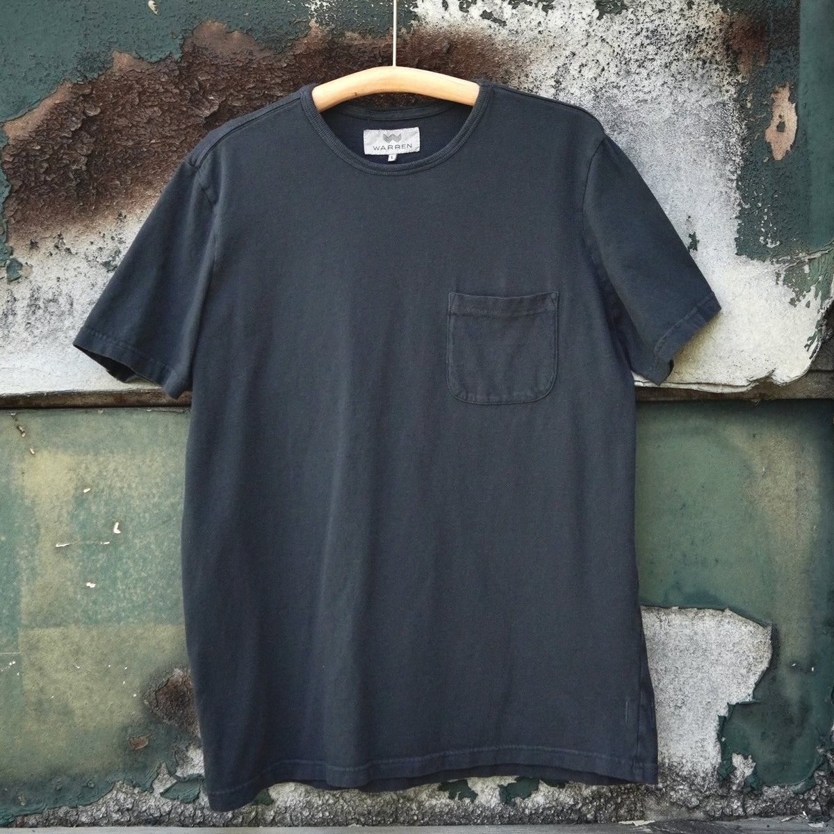 A off black short-sleeve pocket t-shirt hanging on a wooden hanger against a weathered green and white wooden wall.