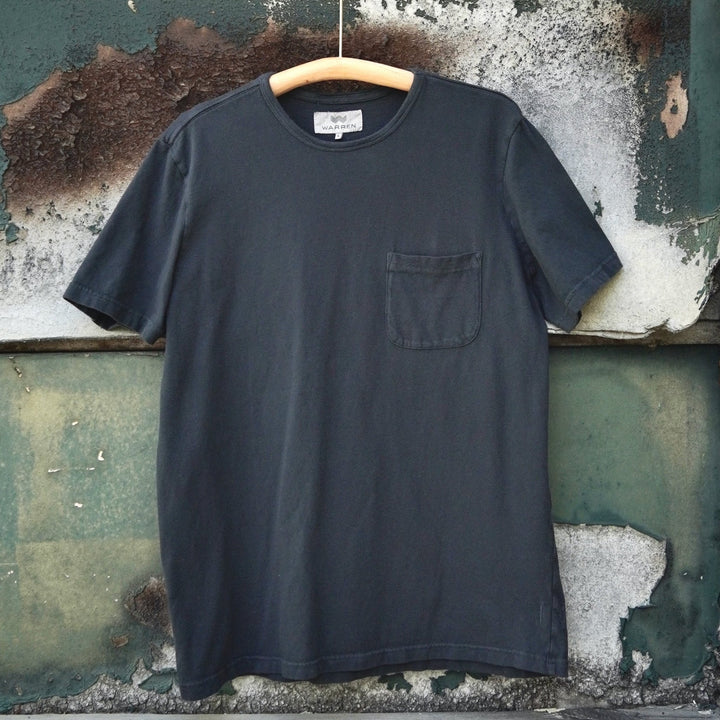 A off black short-sleeve pocket t-shirt hanging on a wooden hanger against a weathered green and white wooden wall.