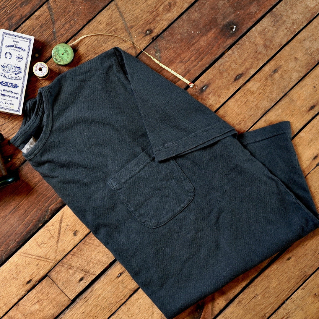 A folded off black cotton t-shirt with a chest pocket, styled on a rustic wooden plank floor alongside vintage sewing accessories including a spool of thread, a small box of buttons, and a wooden spool.