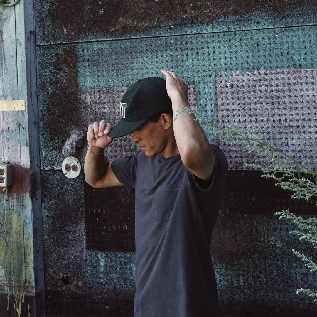 A man wearing a black baseball cap with a white embroidered logo and an off black t-shirt stands in front of a weathered, textured metal wall.