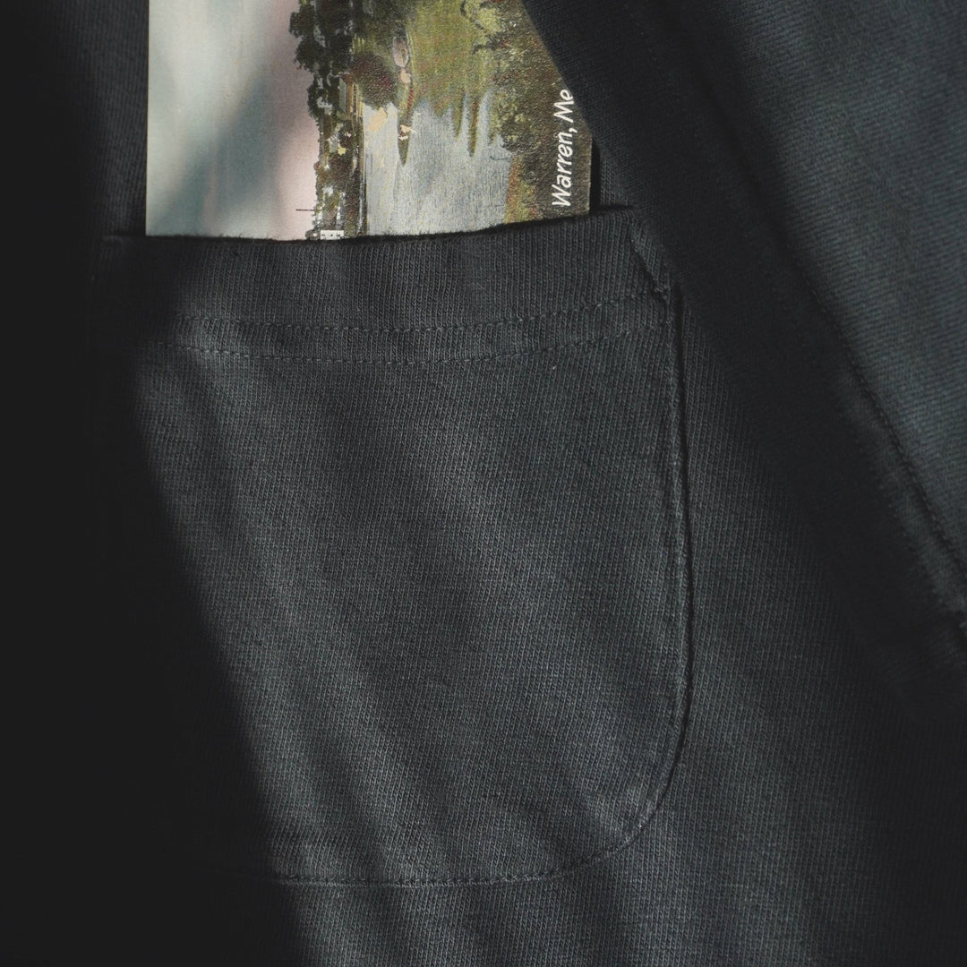 A close-up of an off-black fabric pocket with a small printed postcard or photo card tucked into it. The card shows a landscape scene with trees and water, and the text &