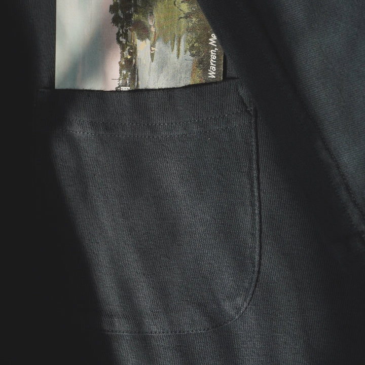 A close-up of an off-black fabric pocket with a small printed postcard or photo card tucked into it. The card shows a landscape scene with trees and water, and the text &