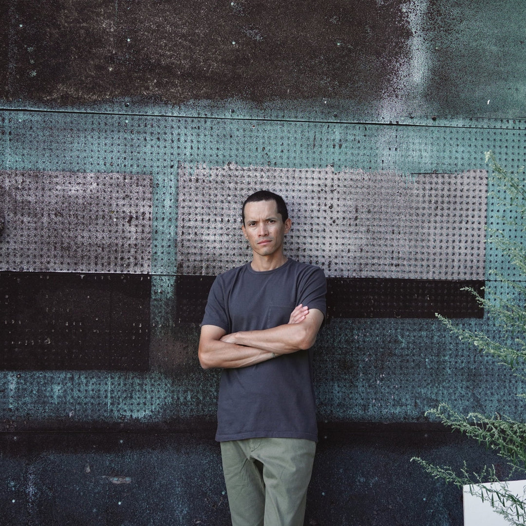 A man with arms crossed stands in front of a weathered teal and black metal wall, wearing an off-black T-shirt and olive green pants.