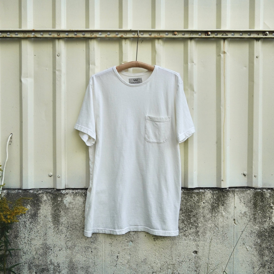 A white short-sleeve crewneck pocket t-shirt hanging on a wooden hanger against a weathered corrugated metal wall.