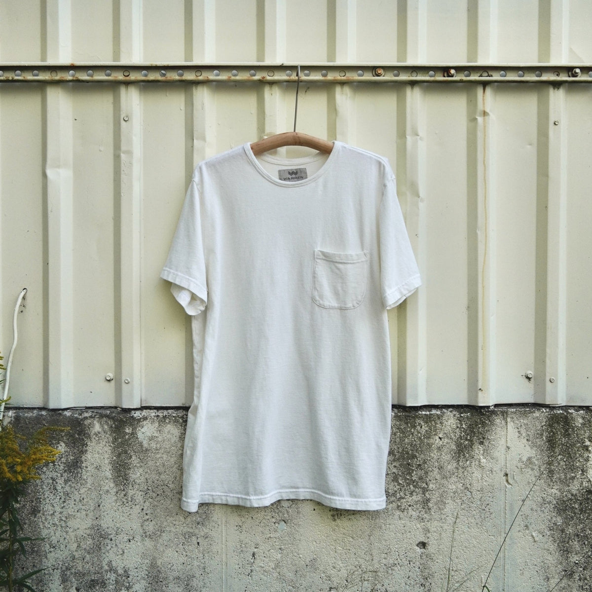 A white short-sleeve crewneck pocket t-shirt hanging on a wooden hanger against a weathered corrugated metal wall.