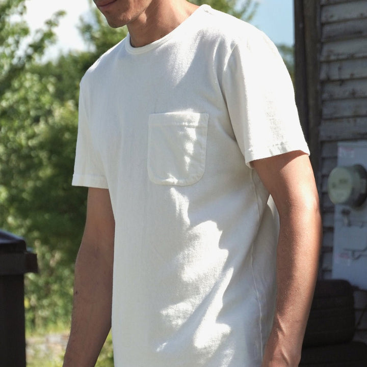 A man wearing a white short-sleeve pocket t-shirt and dark jeans, standing outdoors near a rustic wooden wall and green foliage.