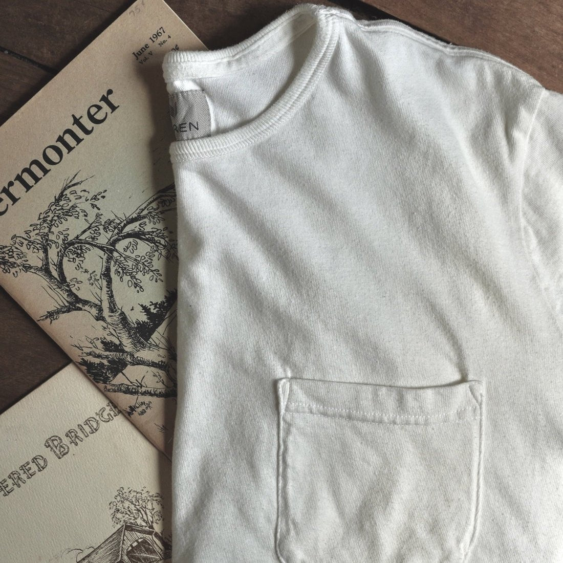 A folded off-white cotton t-shirt with a single front patch pocket, laid flat on a dark wooden surface next to vintage illustrated booklets.