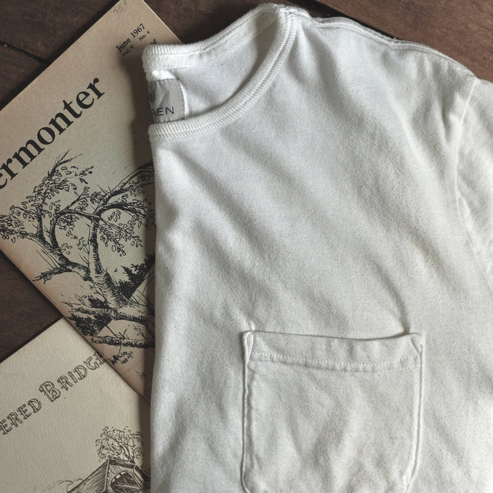 A folded off-white cotton t-shirt with a single front patch pocket, laid flat on a dark wooden surface next to vintage illustrated booklets.