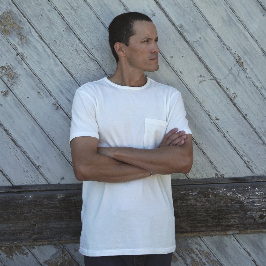 A man wearing a white short-sleeve crewneck t-shirt with a chest pocket, standing with arms crossed against a weathered gray wooden plank wall.