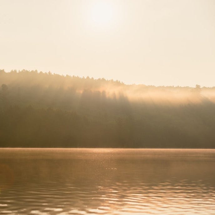 A serene landscape photograph of a calm lake with misty forested hills in the background, illuminated by warm golden sunlight and sun rays filtering through the trees.