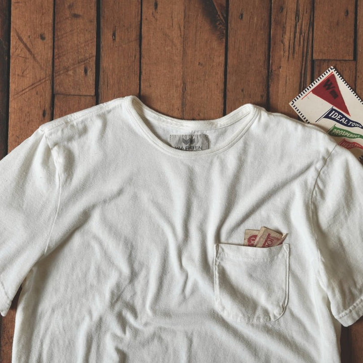 An off-white short-sleeve pocket t-shirt laid flat on a rustic wooden plank background, featuring a small graphic print on the pocket.
