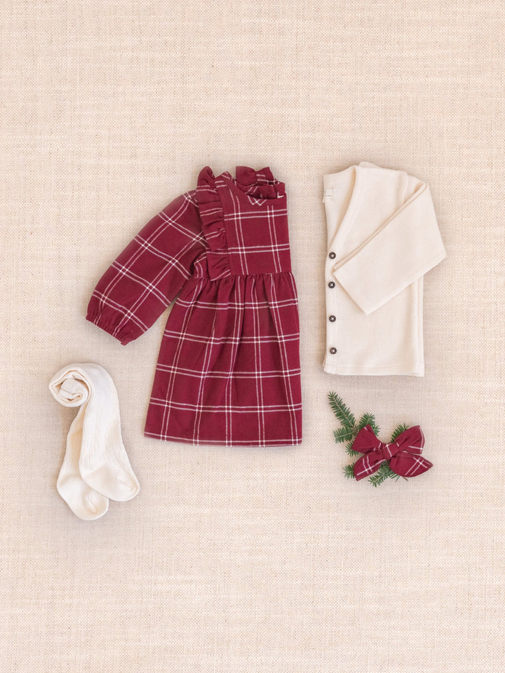 A styled flat-lay of a baby outfit set on a beige linen background, Celyn Flannel Ruffle Long Sleeve Dress, an off-white button-front cardigan, white socks, and a small red bow with green pine sprigs.