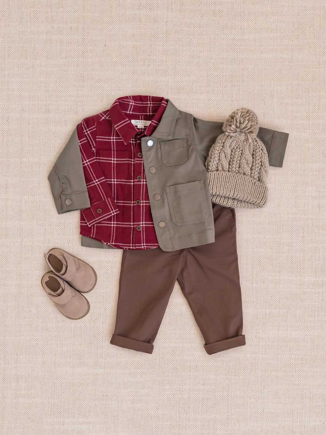 A  three-piece outfit set laid flat on a textured beige fabric background, including a red and white plaid flannel shirt, an olive green button-up jacket, brown pants, a beige cable-knit beanie with a pom-pom, and matching beige slippers.