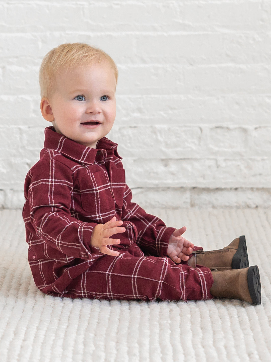 A baby sitting on a white textured blanket in front of a white brick wall, wearing a Colored Organics&