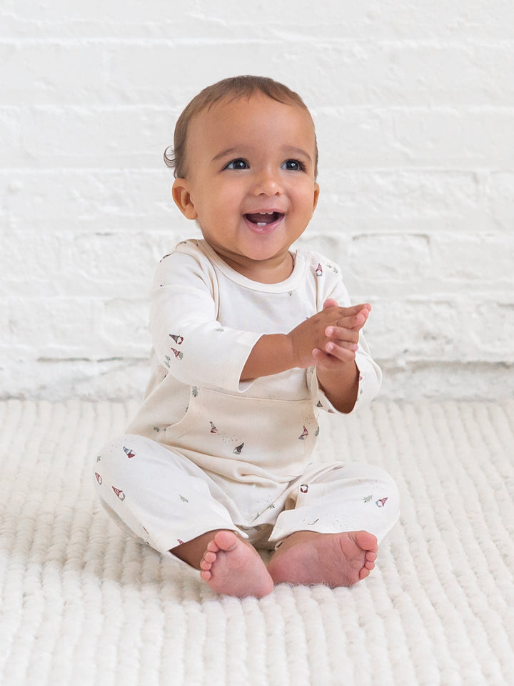 A smiling baby sitting on a textured cream blanket in front of a white brick wall, wearing a Colored Organics&