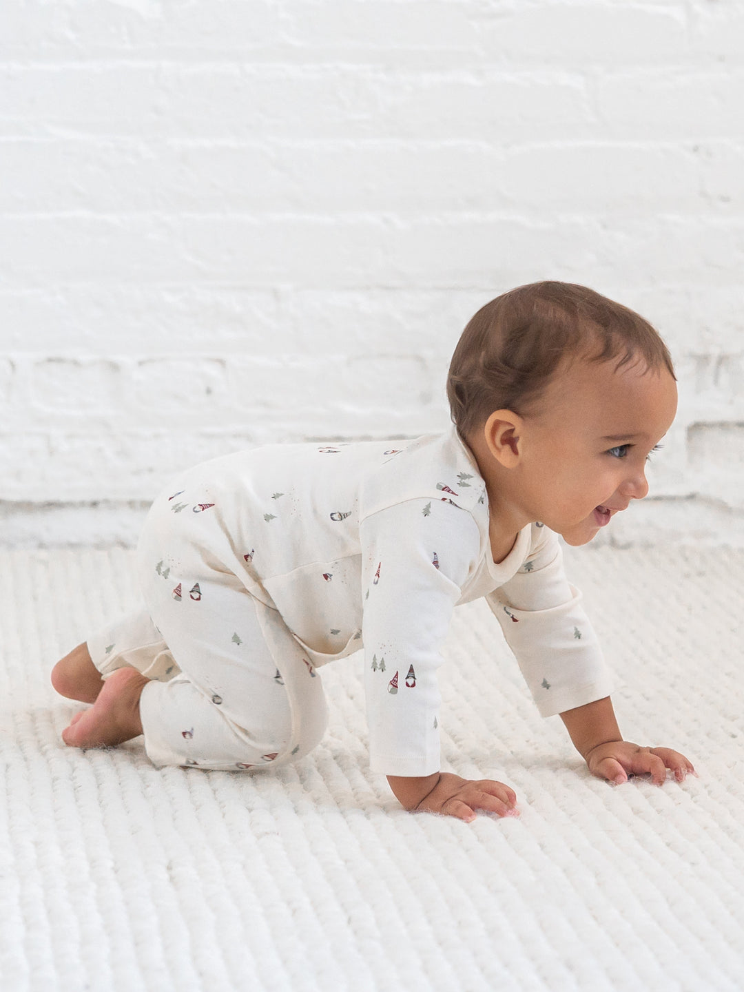 A baby crawling on a white textured blanket in front of a white brick wall, wearing a Una Pocket Romper in gnome print.