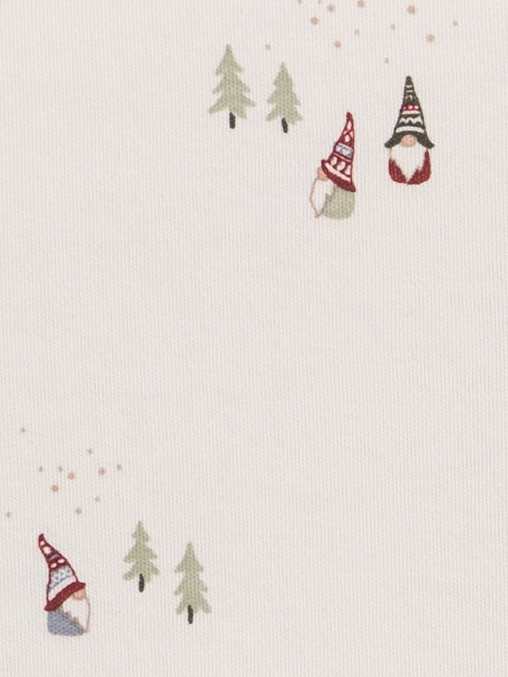 A close-up of a light cream fabric with a delicate printed pattern of small Christmas gnomes wearing red and black hats, green pine trees, and tiny gold speckles.