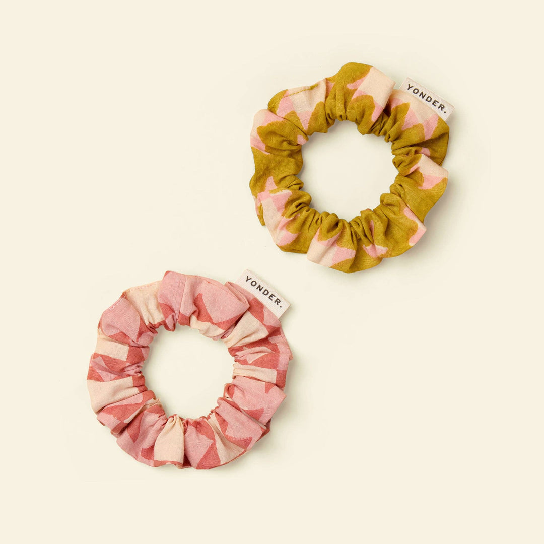 Two fabric hair scrunchies on a warm cream background. One scrunchie is yellow and pink, and the other is pink and beige.