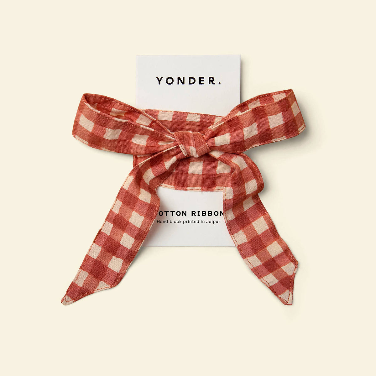 A red and cream gingham cotton ribbon tied in a bow, presented on a white backing card with the brand name 'YONDER.' printed at the top, set against a solid cream background.
