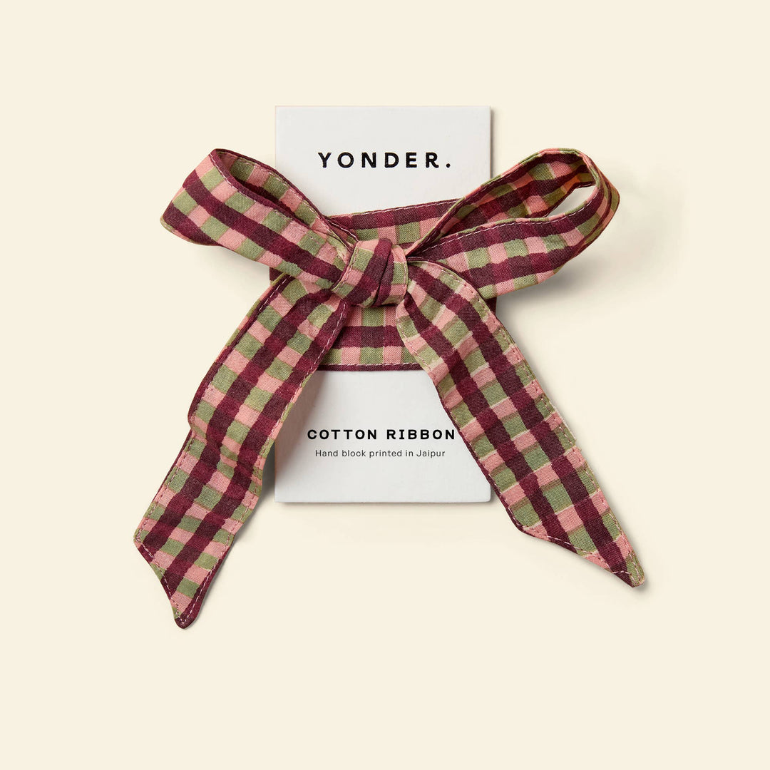 A yonder cotton ribbon tied in a bow around a white card, photographed on a warm cream background. The ribbon has a check pattern in burgundy, dusty pink, and sage green.