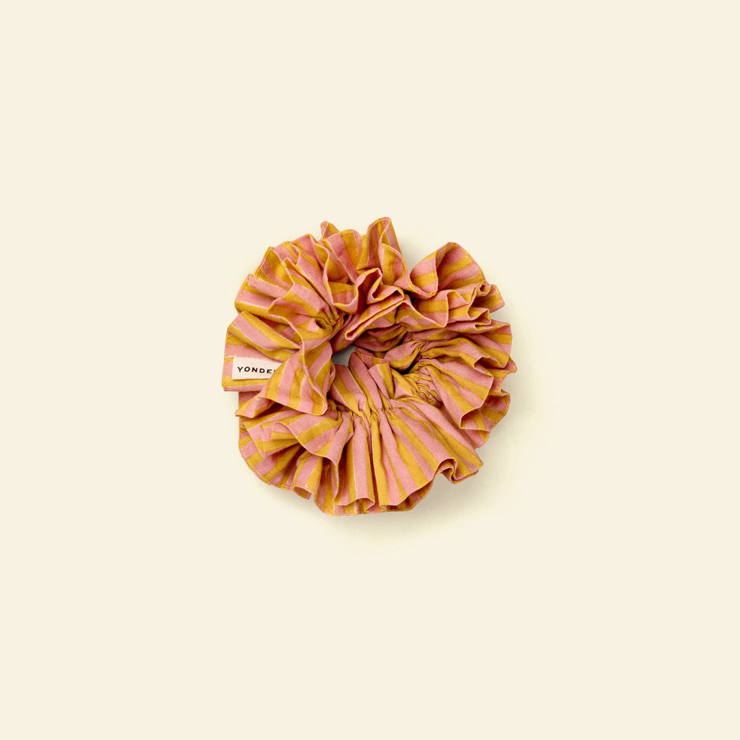 A ruffled fabric hair scrunchie with vertical stripes in pink and mustard yellow, featuring a small white brand tag that reads &